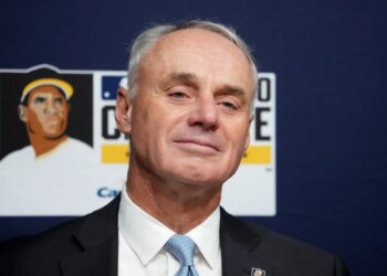 MLB Commissioner Rob Manfred in a suit and tie, against a dark blue background with a baseball illustration.