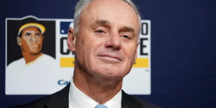 MLB Commissioner Rob Manfred in a suit and tie, against a dark blue background with a baseball illustration.