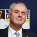 MLB Commissioner Rob Manfred in a suit and tie, against a dark blue background with a baseball illustration.