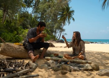 A man and a woman sitting on a beach in front of a makeshift fire pit.