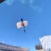 A parachuter with a white and blue parachute caught in a field goal net above a stadium.