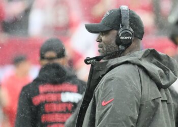 Tampa Bay Buccaneers head coach Todd Bowles wearing a headset and a black cap with a logo, watching from the sideline.