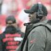 Tampa Bay Buccaneers head coach Todd Bowles wearing a headset and a black cap with a logo, watching from the sideline.