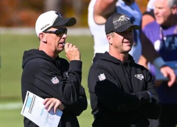 Two football coaches watching a game, one wearing sunglasses and holding a paper.