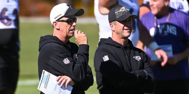 Two football coaches watching a game, one wearing sunglasses and holding a paper.