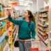 Woman with a grocery cart taking a box of juice from a top shelf in a supermarket.