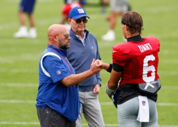 Giants head coach Brian Daboll fist-bumps quarterback Jaxson Dart, with owner John Mara standing behind them, on a football field.