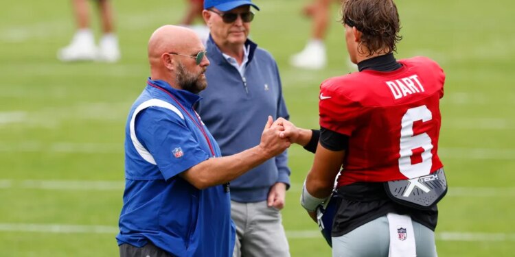 Giants head coach Brian Daboll fist-bumps quarterback Jaxson Dart, with owner John Mara standing behind them, on a football field.