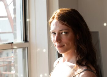 Bree O’Donnell looking at the viewer next to her bedroom window. Sunlight is reflected from a spectrum that bounces little discs of light all over her face and the wall behind her.
