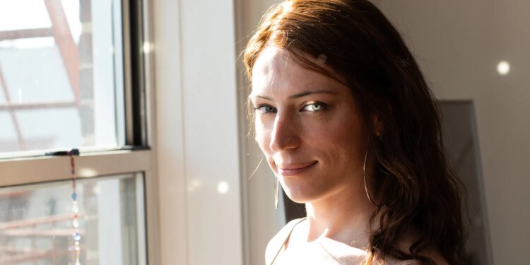 Bree O’Donnell looking at the viewer next to her bedroom window. Sunlight is reflected from a spectrum that bounces little discs of light all over her face and the wall behind her.