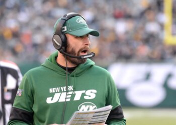 A coach for the New York Jets wearing a green hoodie, green cap, and a headset, with his mouth open, likely shouting instructions during a game.