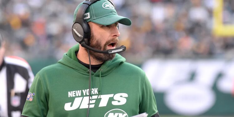 A coach for the New York Jets wearing a green hoodie, green cap, and a headset, with his mouth open, likely shouting instructions during a game.