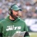 A coach for the New York Jets wearing a green hoodie, green cap, and a headset, with his mouth open, likely shouting instructions during a game.