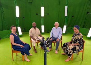 Two men and two women sitting in a semi circle on chairs. The room around the people is covered in green fabric, the floor is also green, and the group are all looking into an array of cameras.