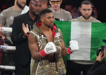 Boxer Anthony Joshua wearing white Everlast gloves and a camouflage vest, with a Nigerian flag behind him.