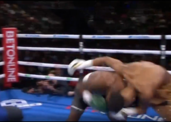 A boxer in black and white shorts falls to the mat while his opponent stands over him.
