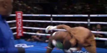 A boxer in black and white shorts falls to the mat while his opponent stands over him.