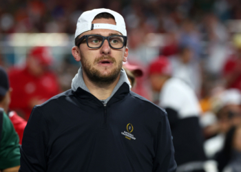 A man wearing a white cap backwards, glasses, and a black jacket with a "National Championship Miami" logo.