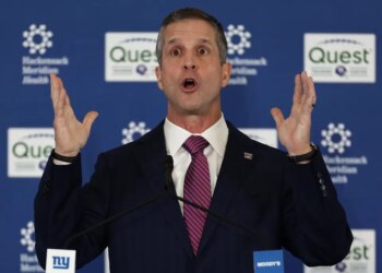New York Giants head coach John Harbaugh speaking at a news conference, with his arms raised and mouth open.