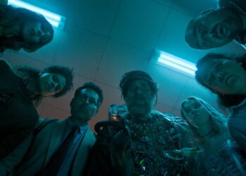 Four woman and three men standing together in a semicircle looking down at something on the floor in confusion.