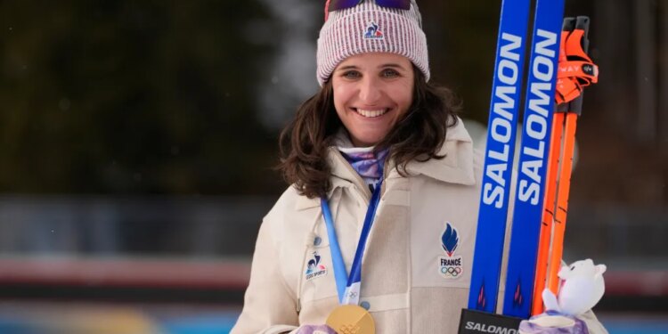 Julia Simon, of France, smiles while holding her gold medal from the women's 15-kilometer individual biathlon race and two pairs of skis.
