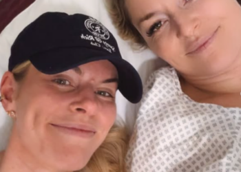 Two women, one in a baseball cap and another in a hospital gown, smile at the camera.