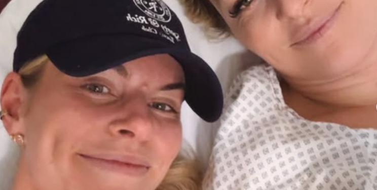 Two women, one in a baseball cap and another in a hospital gown, smile at the camera.