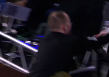 A bald man in a black shirt appears to be falling or climbing over metal scaffolding, his arm outstretched.