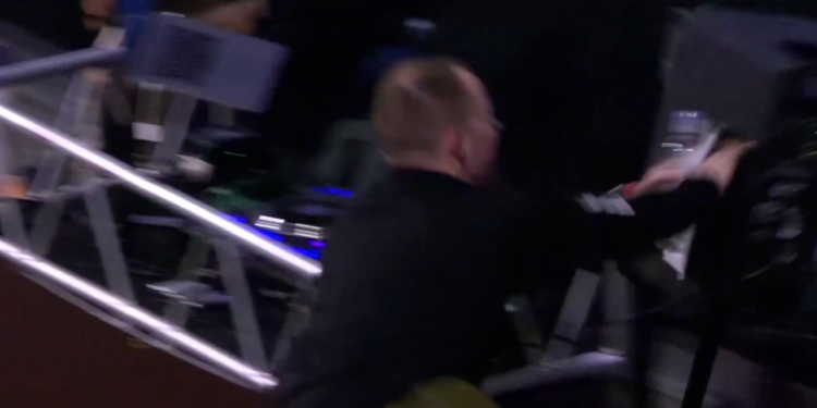 A bald man in a black shirt appears to be falling or climbing over metal scaffolding, his arm outstretched.