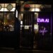 Outside view of a wine bar where you can see people through the window. In neon purple lights is the word EVA AI cafe.