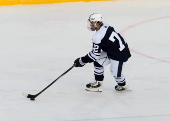 A Penn State hockey player in a navy and white uniform with number 7 on the back and 22 on the sleeve, holding a hockey stick over a puck on the ice.
