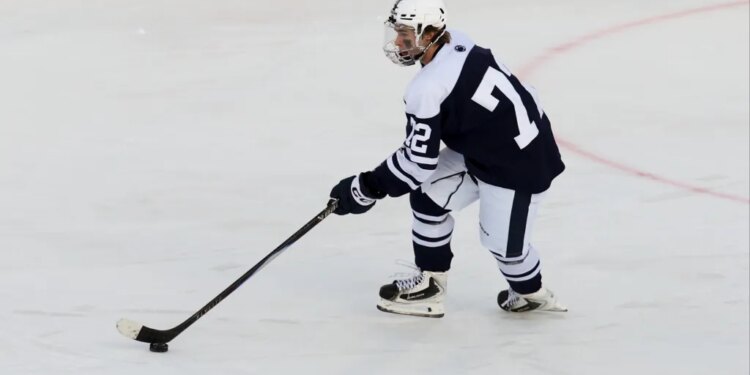 A Penn State hockey player in a navy and white uniform with number 7 on the back and 22 on the sleeve, holding a hockey stick over a puck on the ice.
