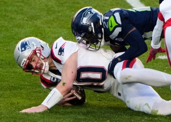 Drake Maye, wearing a white uniform and gray helmet, is sacked by Devon Witherspoon, wearing a dark blue uniform and helmet, during Super Bowl 60.