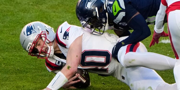 Drake Maye, wearing a white uniform and gray helmet, is sacked by Devon Witherspoon, wearing a dark blue uniform and helmet, during Super Bowl 60.