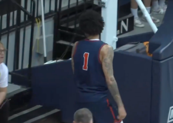 A basketball player in a number 1 jersey stands near the Monmouth logo on a court, while a fan in a white Spider-Man shirt looks on.