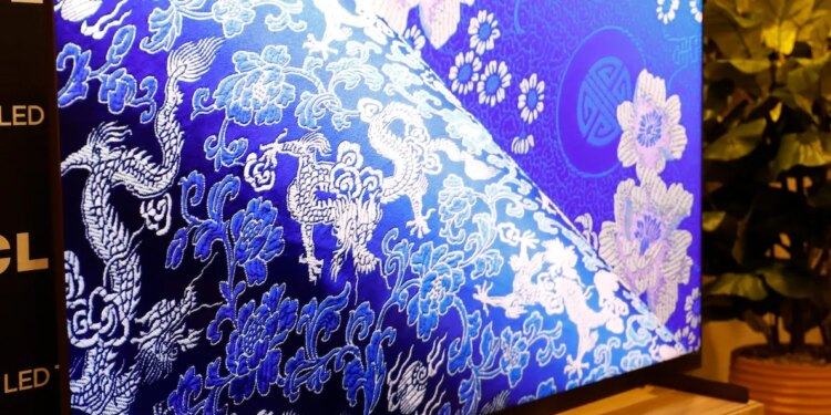 The TCL X11L SQD Mini LED TV on a wooden stand with a TCL-branded backdrop behind it and a potted plant to its side