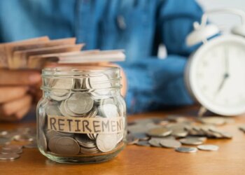 A person putting money into a glass jar labeled "RETIREMENT," with more money and an alarm clock in the background.