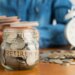 A person putting money into a glass jar labeled "RETIREMENT," with more money and an alarm clock in the background.