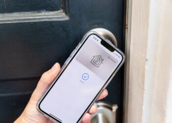 A hand holding an iPhone up to a door. On the phone screen is a picture of a keycard with the Apple Home logo, and the words “My Home” above it. Below it is a blue checkmark in a circle and the word “Done.”