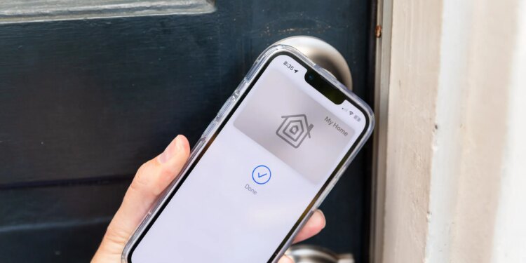 A hand holding an iPhone up to a door. On the phone screen is a picture of a keycard with the Apple Home logo, and the words “My Home” above it. Below it is a blue checkmark in a circle and the word “Done.”