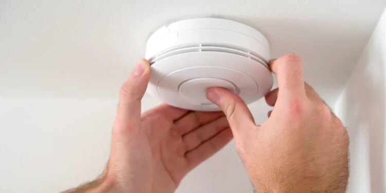 A person installing a white smoke or carbon monoxide detector on a white ceiling.
