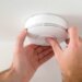 A person installing a white smoke or carbon monoxide detector on a white ceiling.