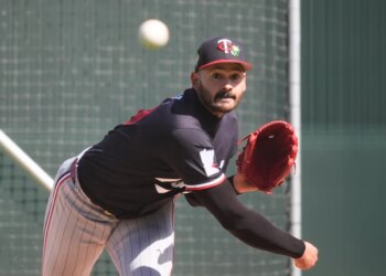 A baseball player in a black Twins cap and black jersey with a red glove winds up to pitch.