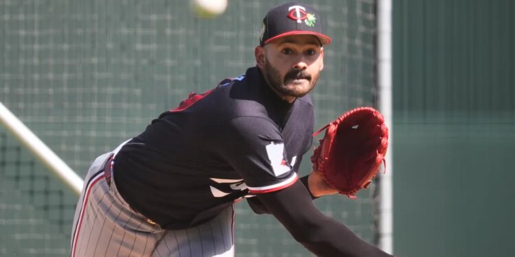 A baseball player in a black Twins cap and black jersey with a red glove winds up to pitch.