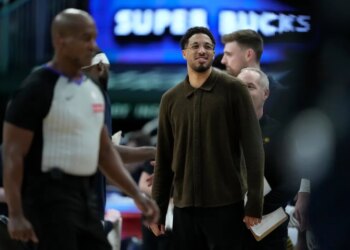 Tyrese Haliburton, wearing a brown collared shirt and glasses, smiling on the sidelines during a game.