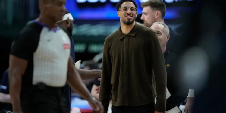 Tyrese Haliburton, wearing a brown collared shirt and glasses, smiling on the sidelines during a game.