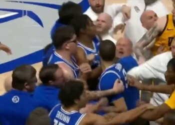 A basketball player in a blue jersey is held back from an opposing player in a yellow jersey, while coaches and other players attempt to de-escalate the situation.