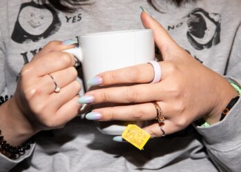 Person in a gray sweater and pastel nails holding a white tea mug while wearing a pink ceramic Oura Ring 4.