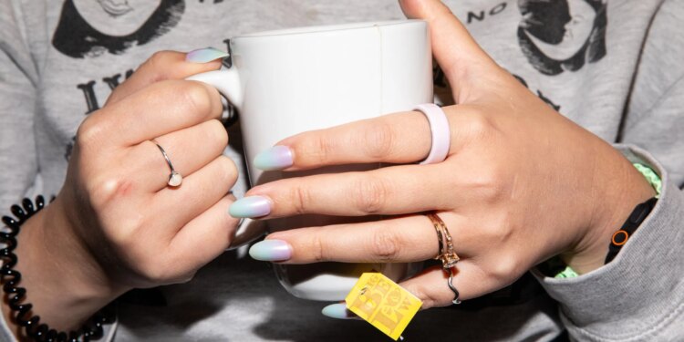 Person in a gray sweater and pastel nails holding a white tea mug while wearing a pink ceramic Oura Ring 4.
