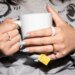 Person in a gray sweater and pastel nails holding a white tea mug while wearing a pink ceramic Oura Ring 4.